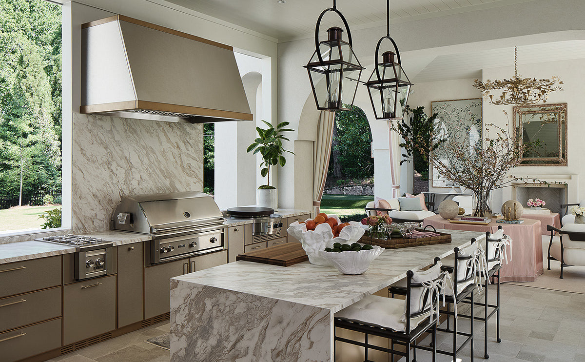 A luxury outdoor kitchen setup featuring a large marble-topped island, stainless steel grill and range hood, beige cabinetry, and black hanging lantern pendant lights in a bright, modern patio setting.