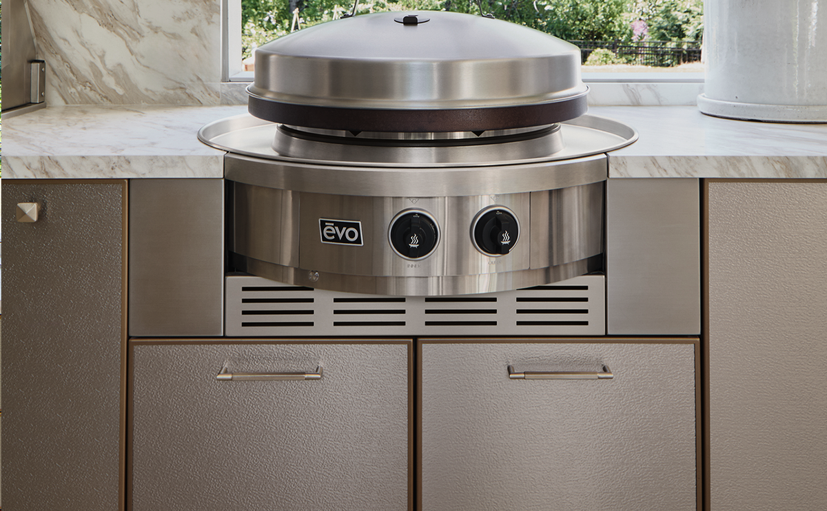 A stainless steel outdoor built-in grill with a domed lid and two front control knobs, installed between beige textured kitchen cabinets on a marble countertop.