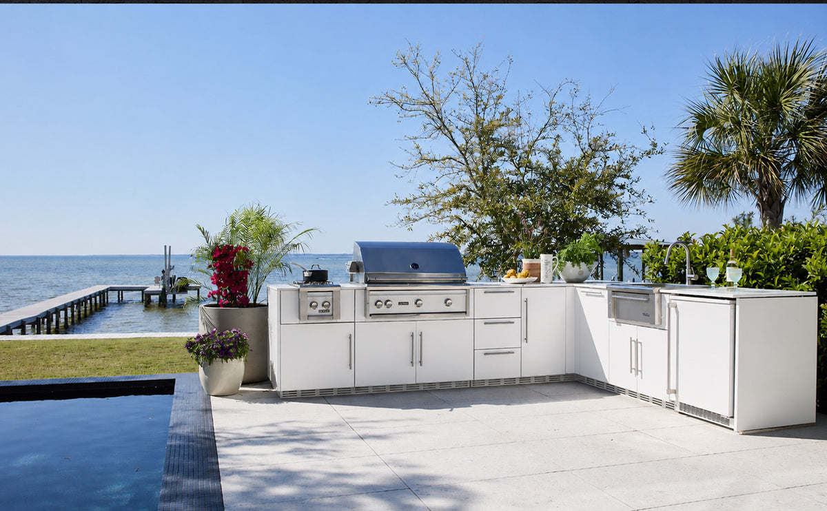 A white outdoor kitchen island set with a stainless steel gas grill, sink, and refrigerator, situated on a concrete patio with a pool, ocean, and palm trees in the background.
