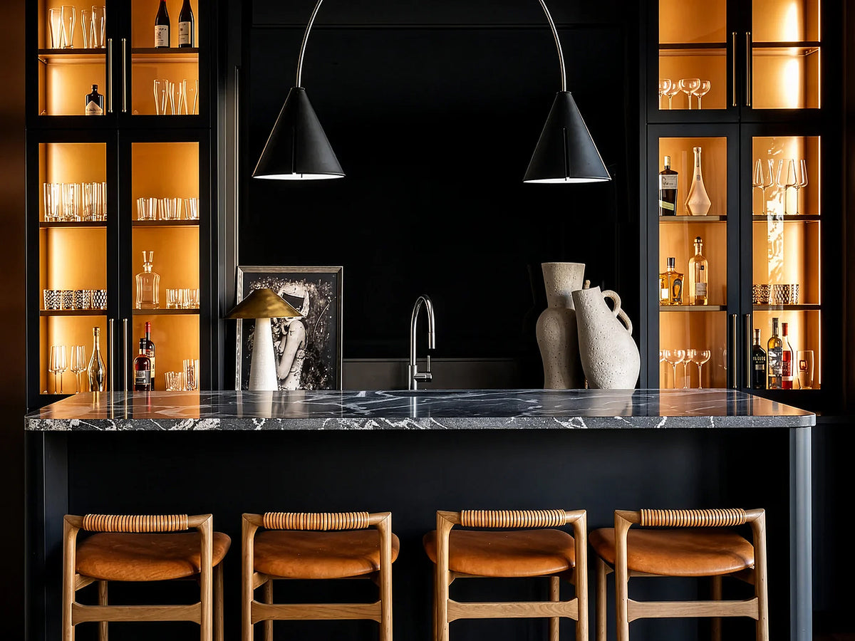 A modern kitchen island with a black marble countertop, a central faucet, decorative vases, and four wooden bar stools with tan leather seats. The background features black cabinetry with warm orange lighting and glassware.