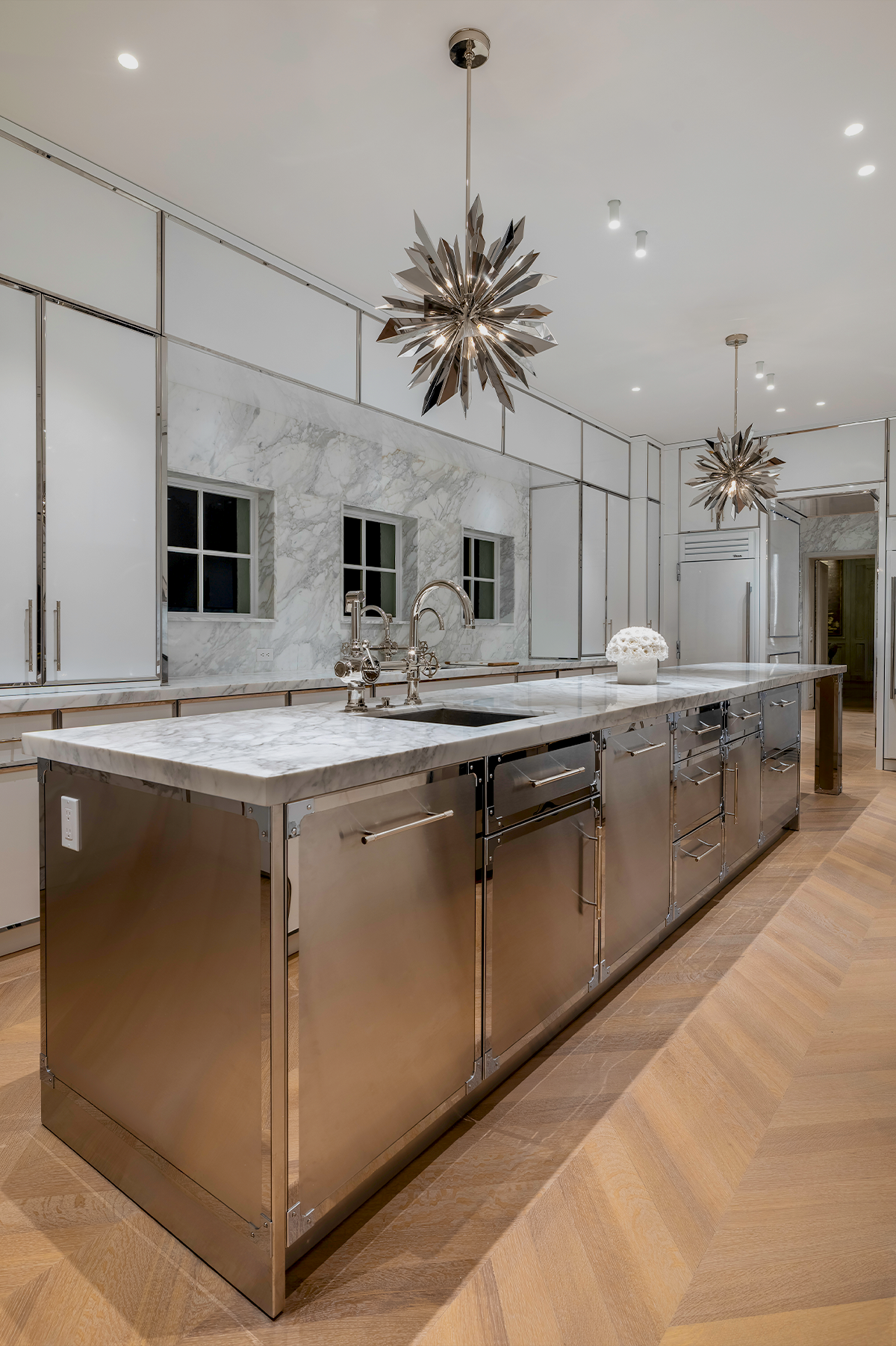 A modern kitchen island with brushed stainless steel cabinets and a white marble countertop, set in a bright kitchen with wood herringbone flooring and starburst pendant lights.