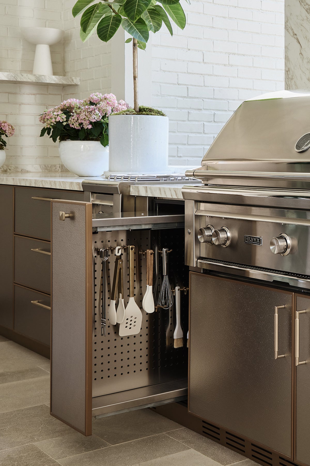 A modern kitchen interior showing a pull-out under-counter cabinet with utensils hanging on a pegboard, next to a stainless steel gas grill. The cabinetry is in a warm taupe-brown finish with brushed brass handles, set against a white brick backsplash and marble countertop.